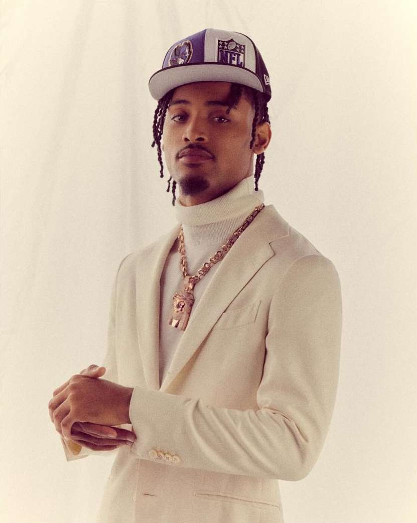 Portrait of an adult male with a baseball cap, wearing a suit and a gold chain necklace in a photo studio with neutral backdrop.