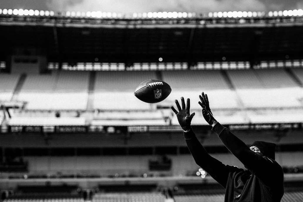 Black and white photo of football in mid-air at stadium, person raises hands to catch it, stadium seating in background.
