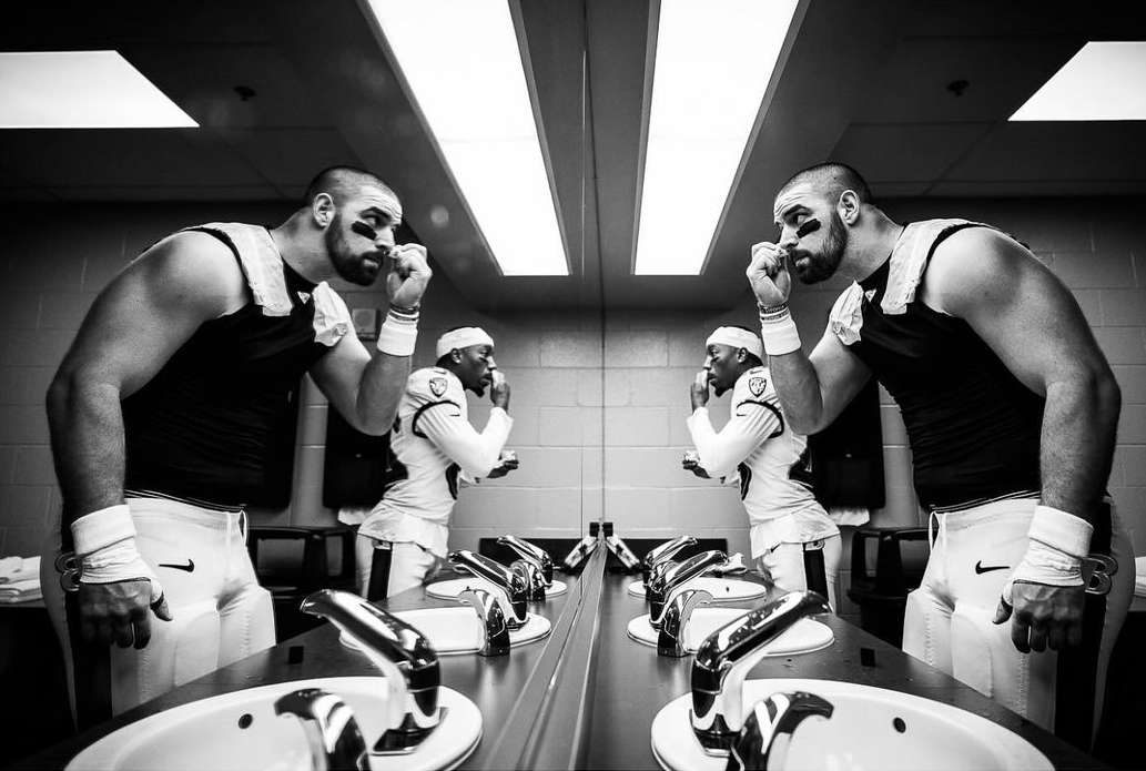Two football players apply eye black in a locker room mirror, reflected with sinks and harsh lighting in a black and white shot.