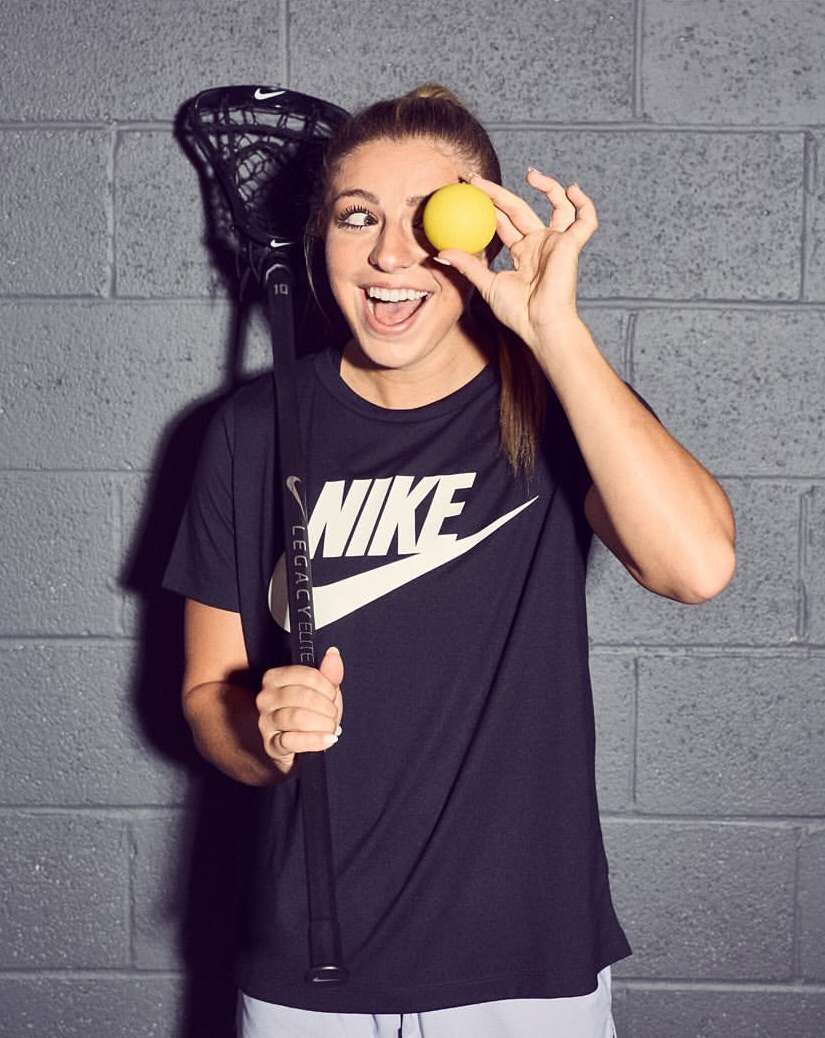 Smiling woman with a lacrosse stick and ball against a gray brick wall wearing a black Nike t-shirt with an open mouth expression.