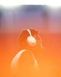Profile portrait of a black man wearing white over-ear headphones and a headband against a warm gradient background.