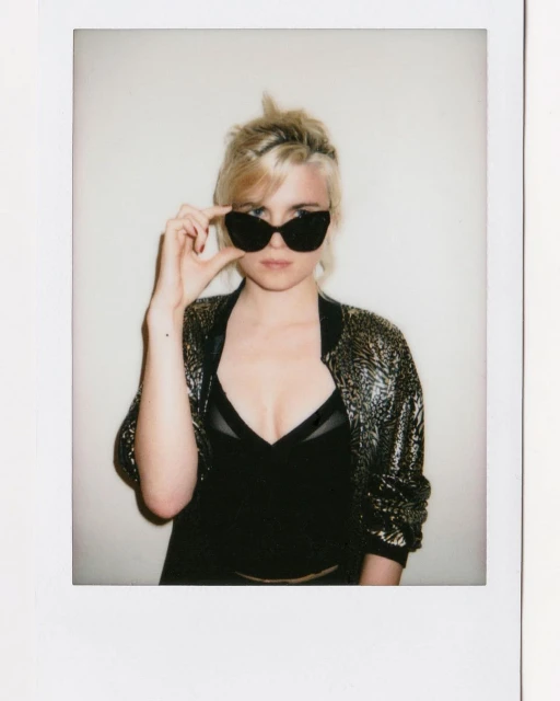 Blonde woman poses in sunglasses and a black top with a gold bomber jacket against a white backdrop in a fashion photo.