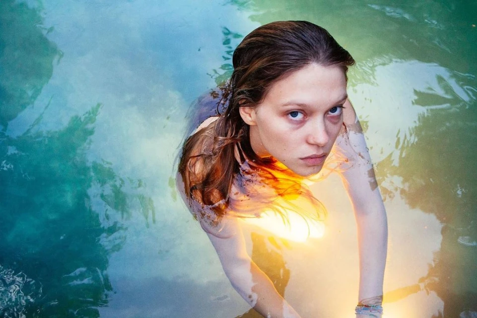 Woman in water gazes up at the camera with a serene expression, the water reflecting the sky's blue and green hues around her.
