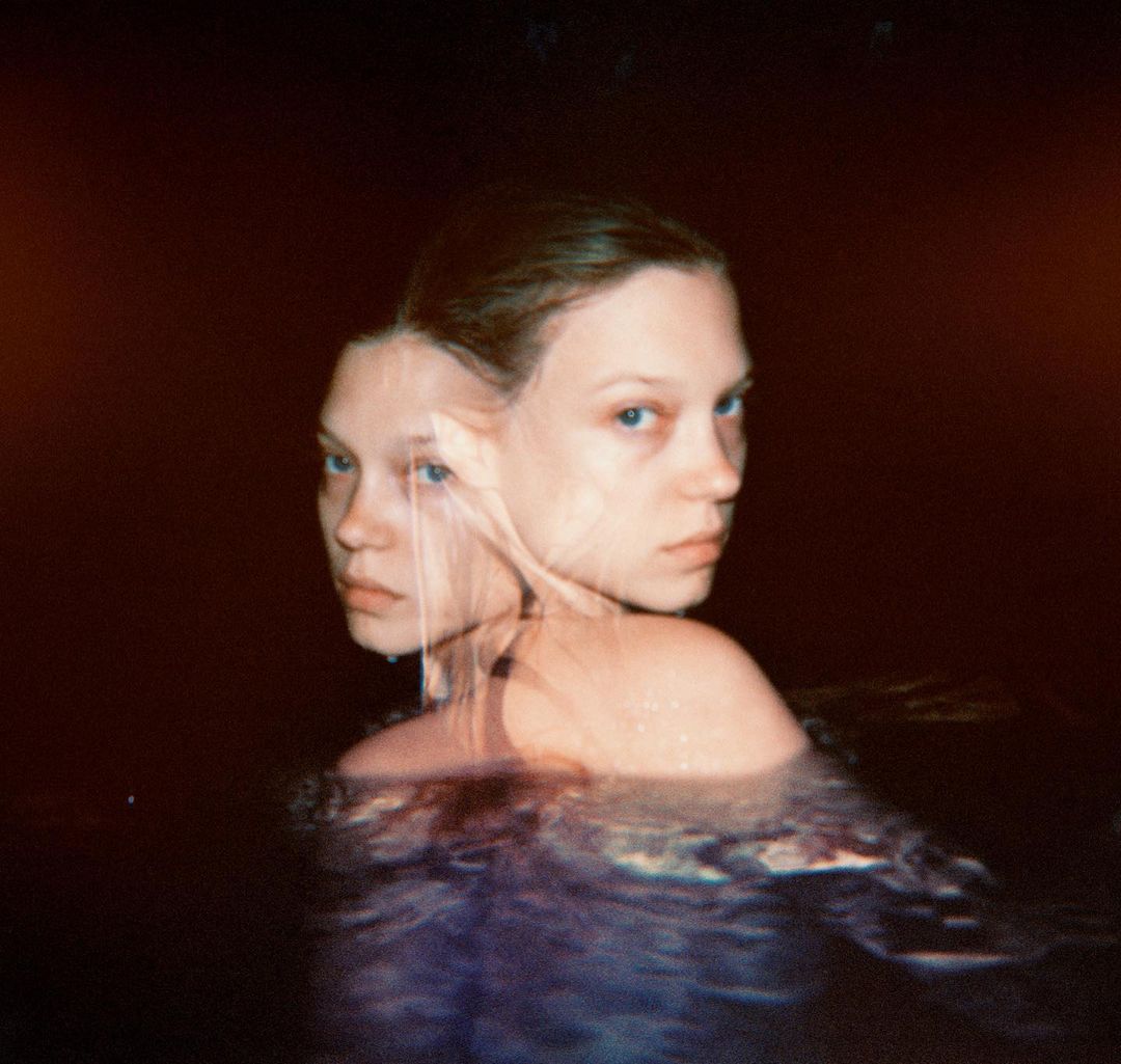 A woman with a serious expression stands waist-deep in water in a dark, moody environment, her face partially obscured by distortion.