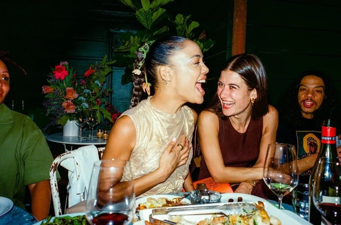 Two women laughing at a table at night. They are at a restaurant and are sitting near food and wine glasses.