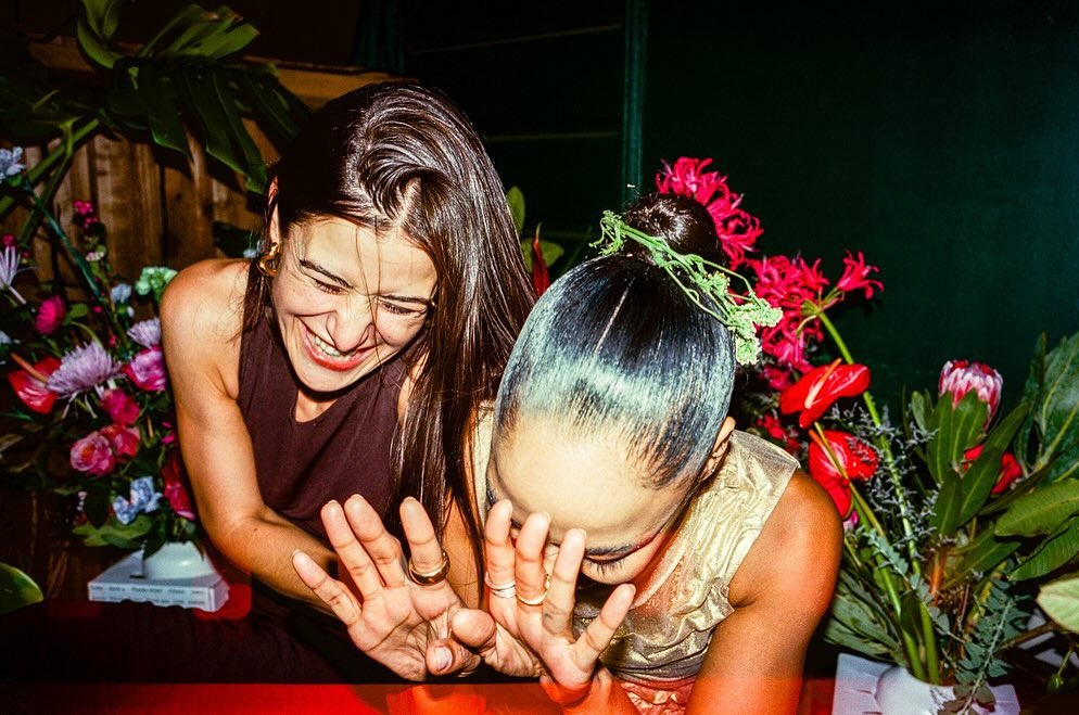 Two women laughing candidly amidst floral arrangements, with one holding her hands up playfully toward the camera.