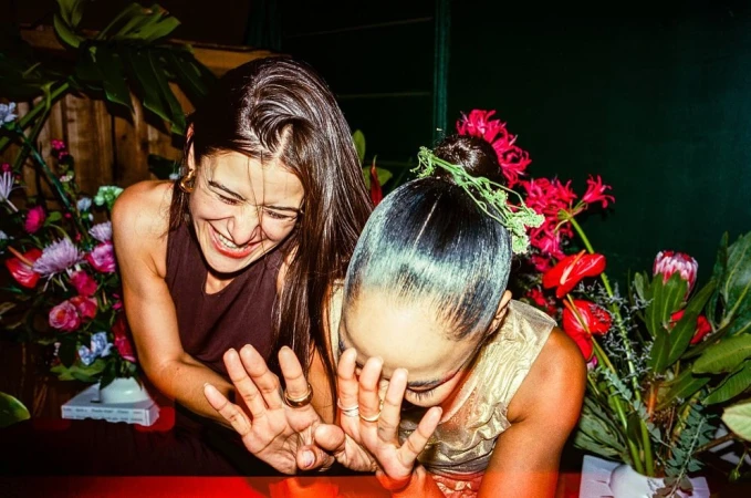 Two women laughing candidly amidst floral arrangements, with one holding her hands up playfully toward the camera.