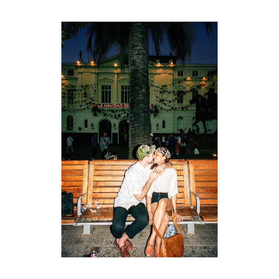 A couple shares a kiss on a wooden bench in an urban environment at night, with a building, palm tree, and streetlights in the background.