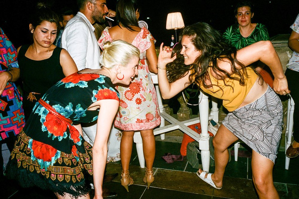 Candid shot of a diverse group of adults interacting at an indoor party, illuminated by lamp light and captured with a wide-angle lens.