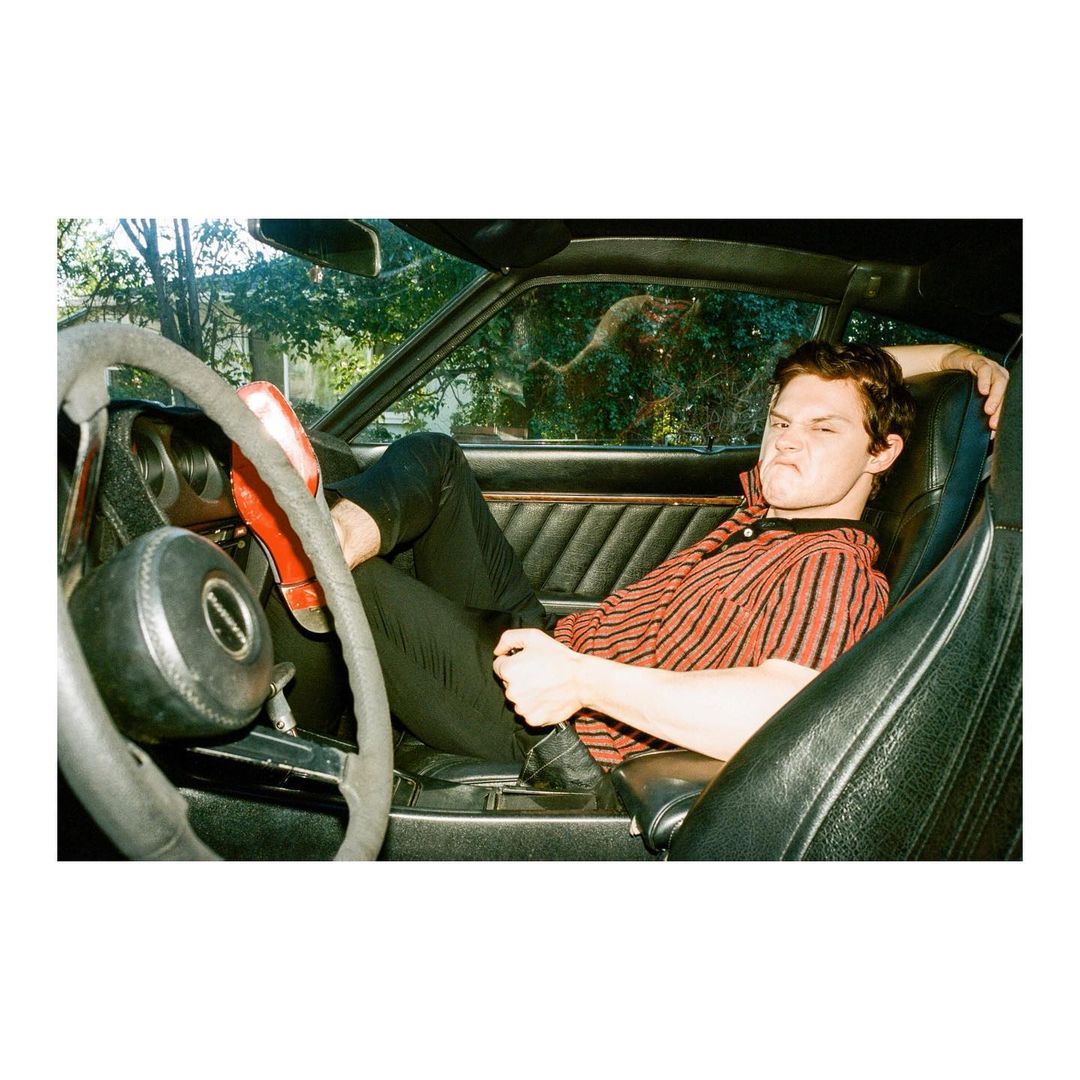 Adult male posing inside a car with red shoes and black pants. He is wearing a red and black stripped shirt. Background includes trees.