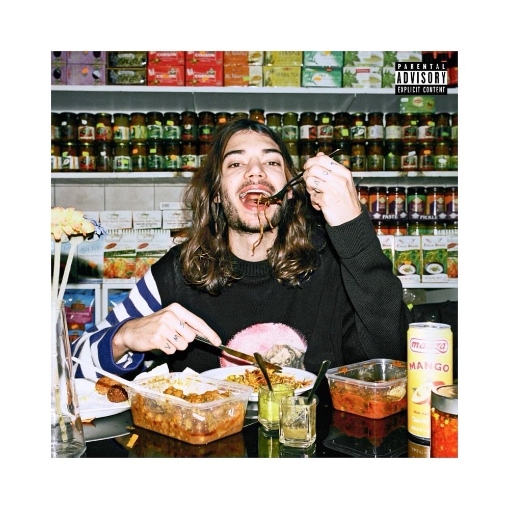 Young adult eats noodles with utensils at a table in front of a shelf full of food containers in an indoor setting with natural lighting.