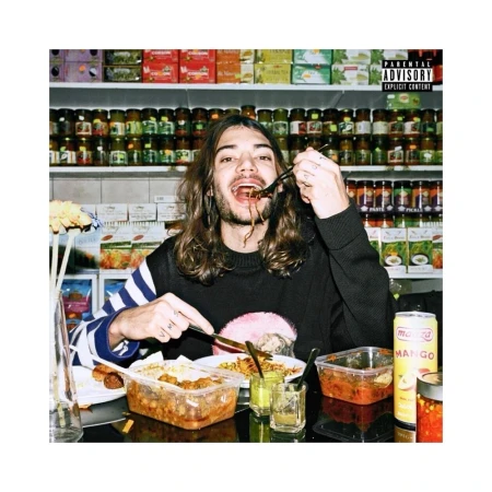 Young adult eats noodles with utensils at a table in front of a shelf full of food containers in an indoor setting with natural lighting.