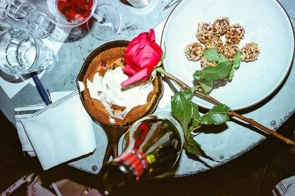 Overhead shot of a dessert in a skillet with a rose, bottle, and wine glass on a textured gray surface, creating an intimate setting.