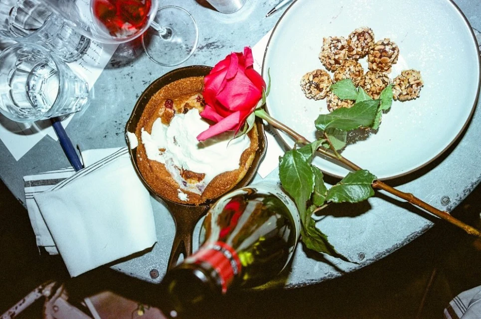 Overhead shot of a dessert in a skillet with a rose, bottle, and wine glass on a textured gray surface, creating an intimate setting.