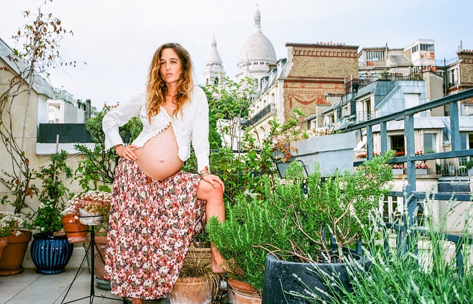 Pregnant woman with floral skirt posing on Paris rooftop terrace surrounded by plants with Sacré-Cœur Basilica in the background.