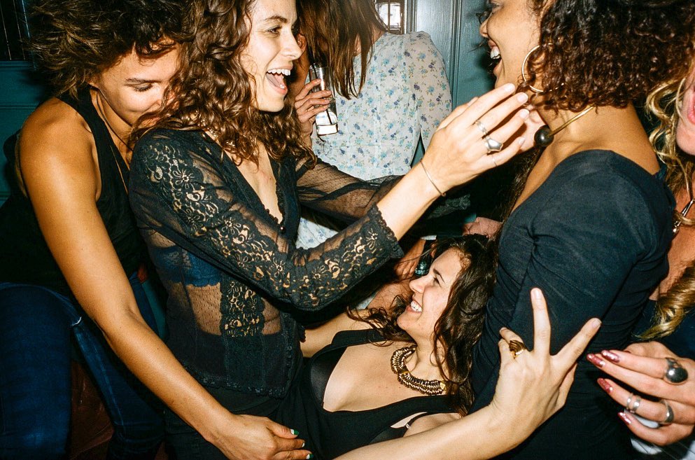 A group of women are gathered indoors, laughing and interacting, in an informal setting with dim lighting, with women wearing black tops.