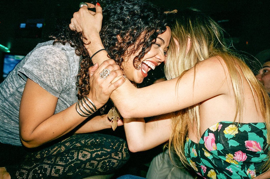 Two women joyfully embrace indoors, with one laughing, showcasing their bond with visible jewelry and casual attire in a dimly lit space.