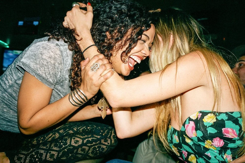 Two women joyfully embrace indoors, with one laughing, showcasing their bond with visible jewelry and casual attire in a dimly lit space.