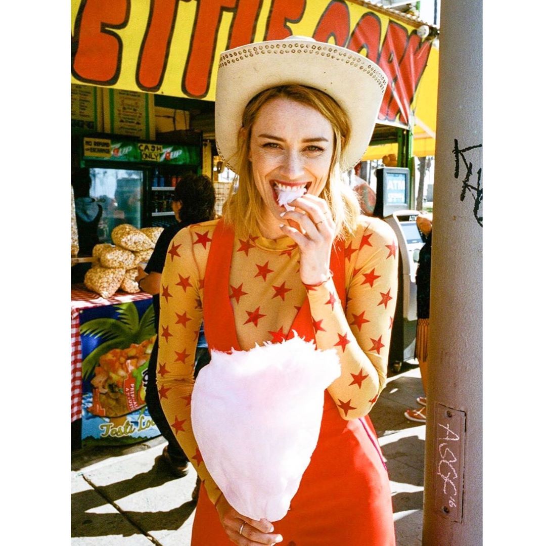 Smiling woman in cowboy hat eating cotton candy at an outdoor fair with a food stand in the background on a sunny day.