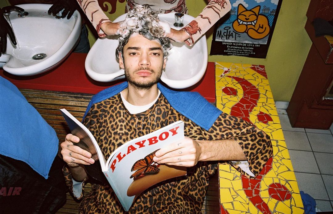Man in a leopard print shirt reads Playboy magazine while getting his hair washed in a salon; poster and colorful mosaic table.