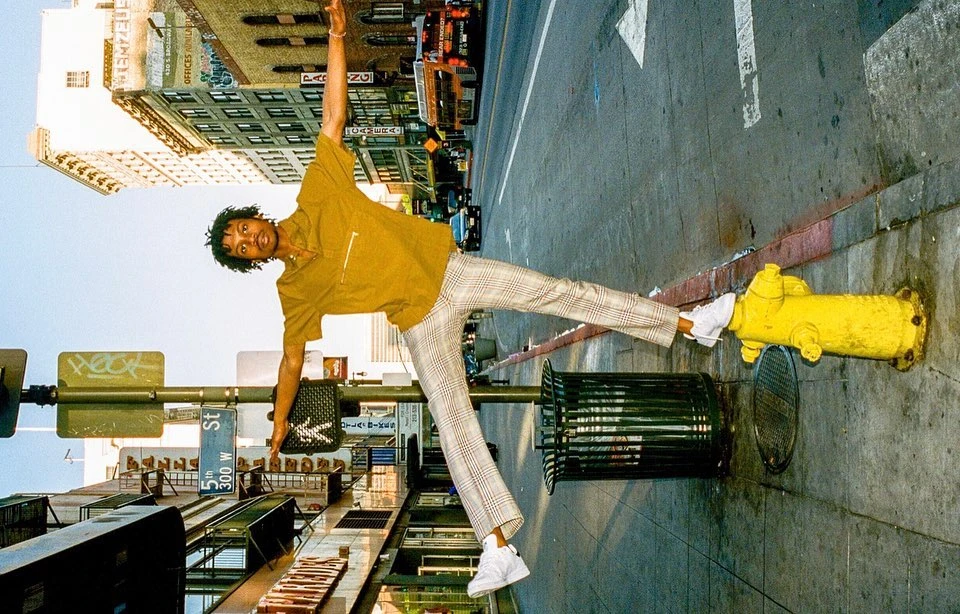 A man balances on a trash can and fire hydrant on a city street, wearing an orange shirt and plaid pants, with buildings in the background.