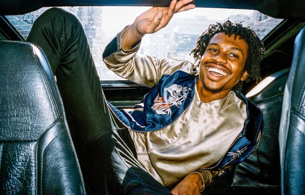Smiling African American man in gold jacket waves from the back seat of a car. Urban environment, daylight, casual fashion.