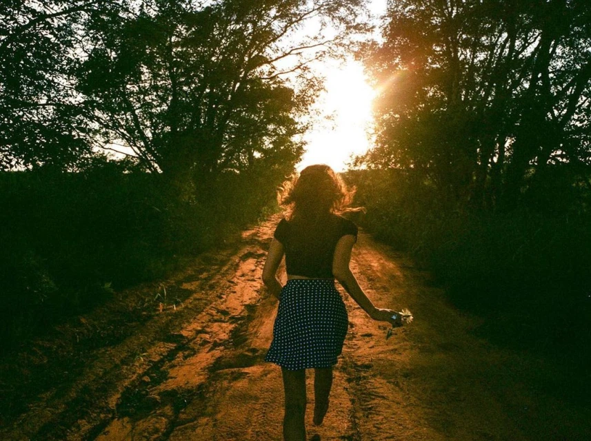 A young woman runs barefoot on a dirt path away from the camera during sunset, trees lining the path. Happy mood, nature scene.