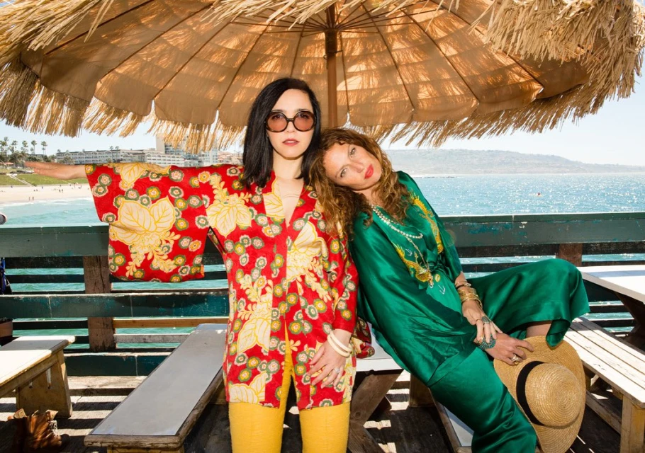 Two women pose on a pier with an umbrella above, wearing colorful patterned outfits with the ocean and coastline in the background.