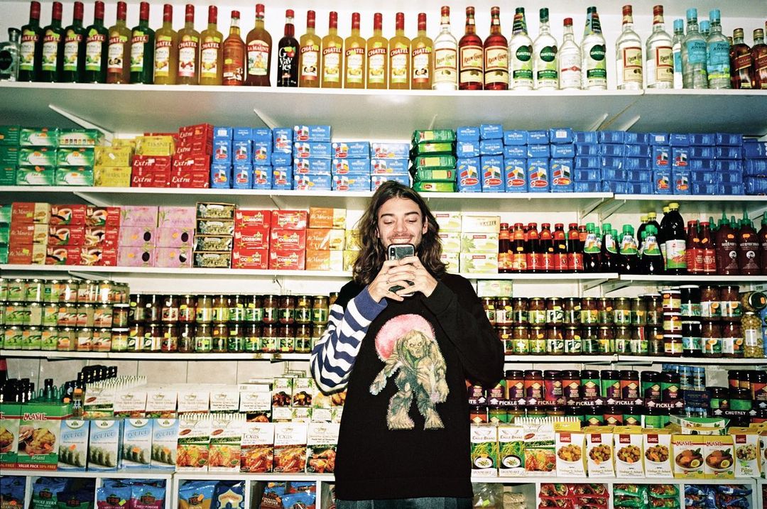 Man using a smartphone standing in front of shelves stocked with grocery and liquor products inside a retail environment.