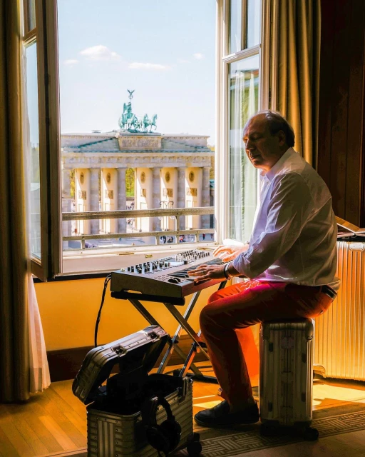 Adult man playing keyboard in an indoor setting by a window with Berlin's Brandenburg Gate in the background on a sunny day.