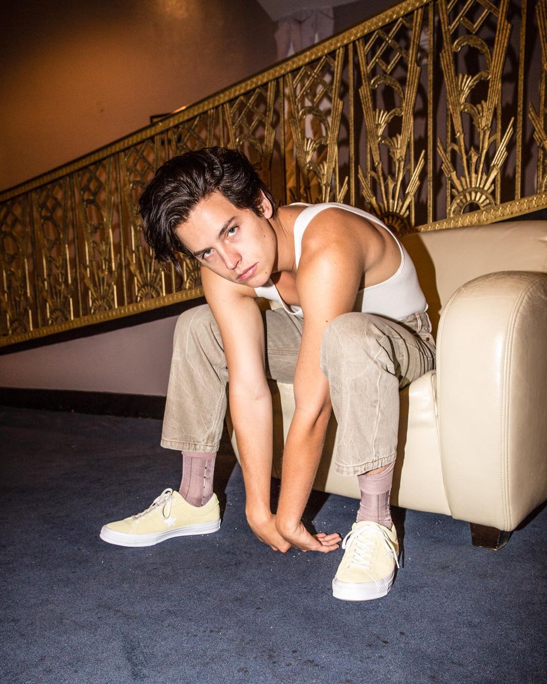 Portrait of a young adult man sitting in a white armchair with a serious expression and wearing casual clothing in an indoor setting.