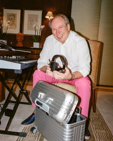 Hans Zimmer holding headphones, seated in a hotel room wearing a white shirt and pink trousers with a metal suitcase in front of him.
