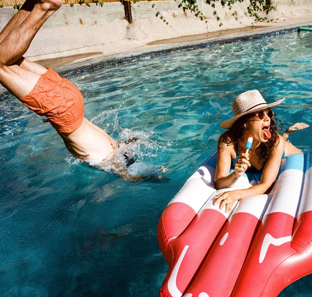 Woman lounges on a red, white, and blue pool float with an ice pop as a man dives into the water of an outdoor swimming pool.