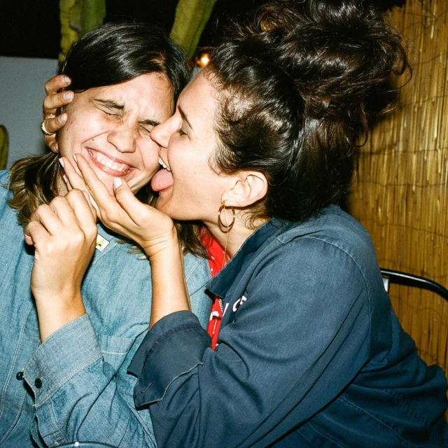 Two adult women in casual denim and dark-blue jackets smiling indoors with cacti in the background. One is sticking her tongue out.