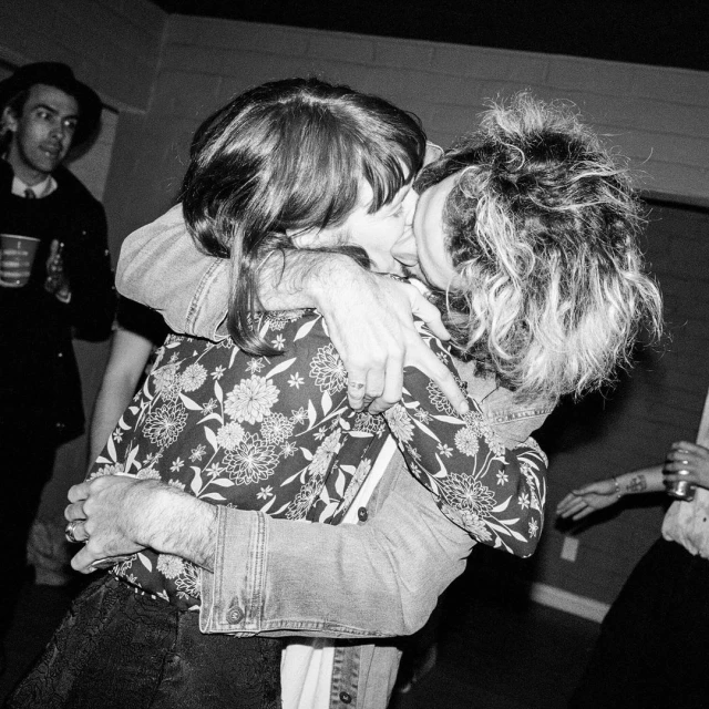 Monochrome photo of a couple embracing and kissing indoors, with two other people visible in the background in soft, diffused lighting.