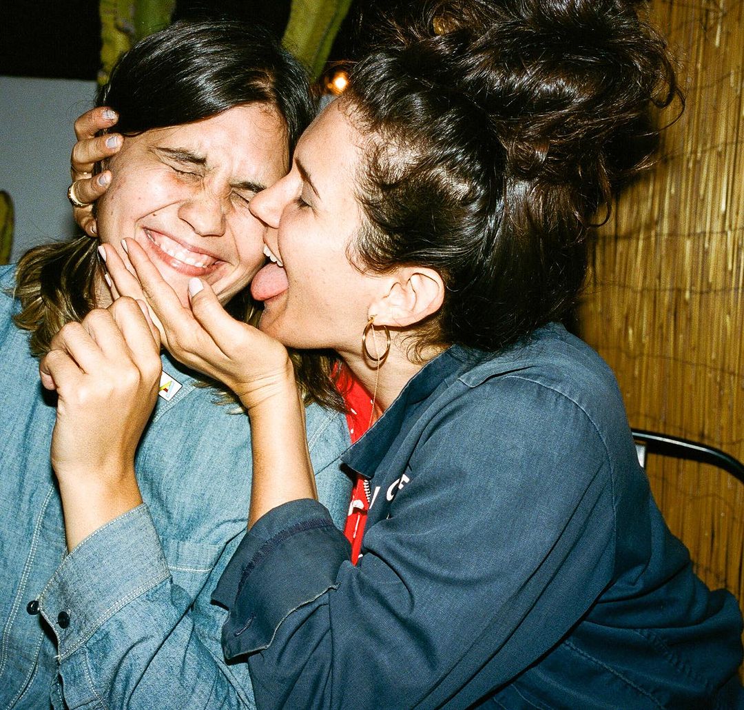Two young adult women pose indoors, smiling and laughing. One woman sticks her tongue out towards the other's face in a playful moment.