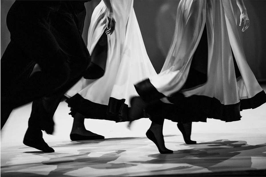 Monochromatic shot of dancers with blurred motions in flowing skirts and dark tights performing on a stage with shadows.