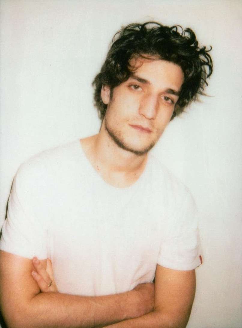 Portrait of a young adult male in a white t-shirt with arms crossed, against a light background in an indoor setting with natural light.
