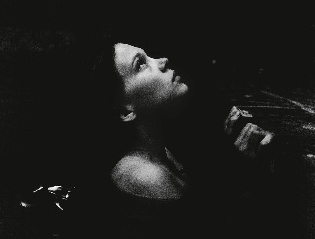 Monochromatic portrait of an adult woman looking up with an open expression. Dark, shallow focus with visible grain, and some visible water.