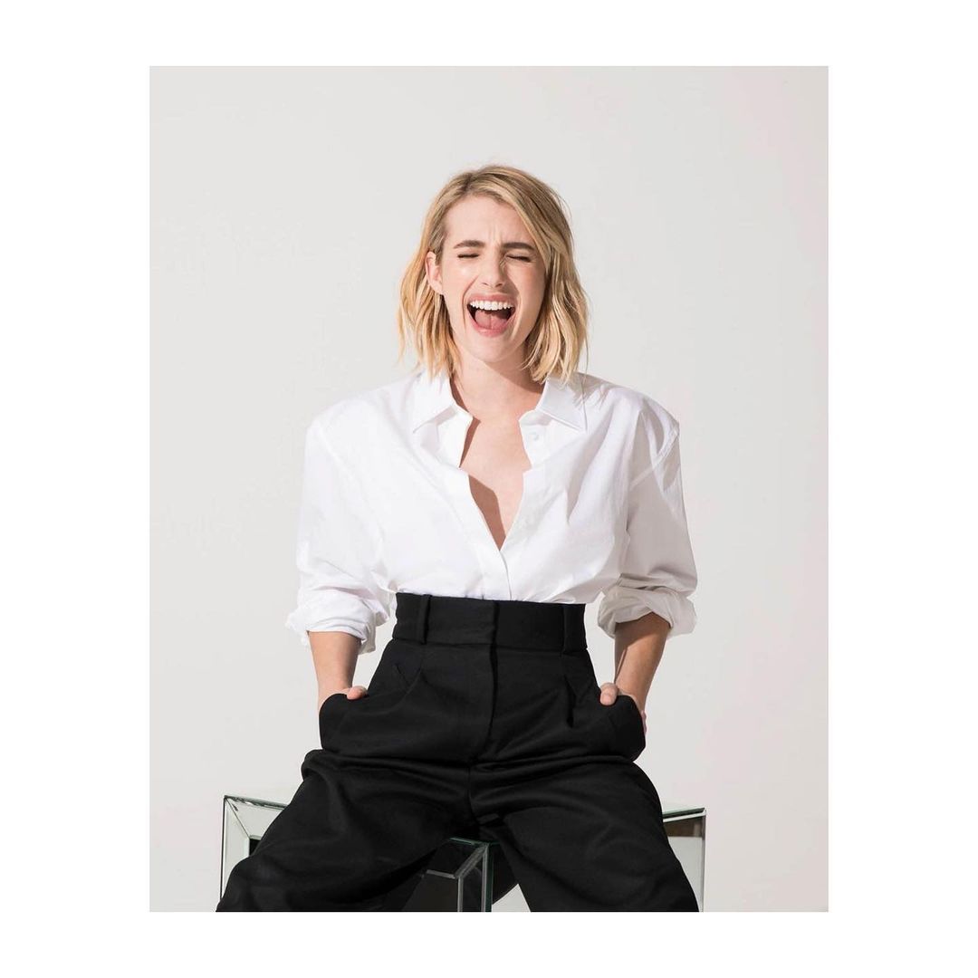 Emma Roberts smiles wide with hands in the pockets of her high-waisted black pants, paired with a white shirt. Sitting on mirror box.