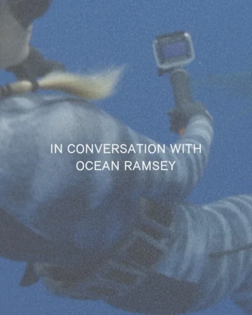 Underwater shot of a person wearing a wetsuit and holding a camera, possibly diving or exploring marine life in a blue aquatic setting.