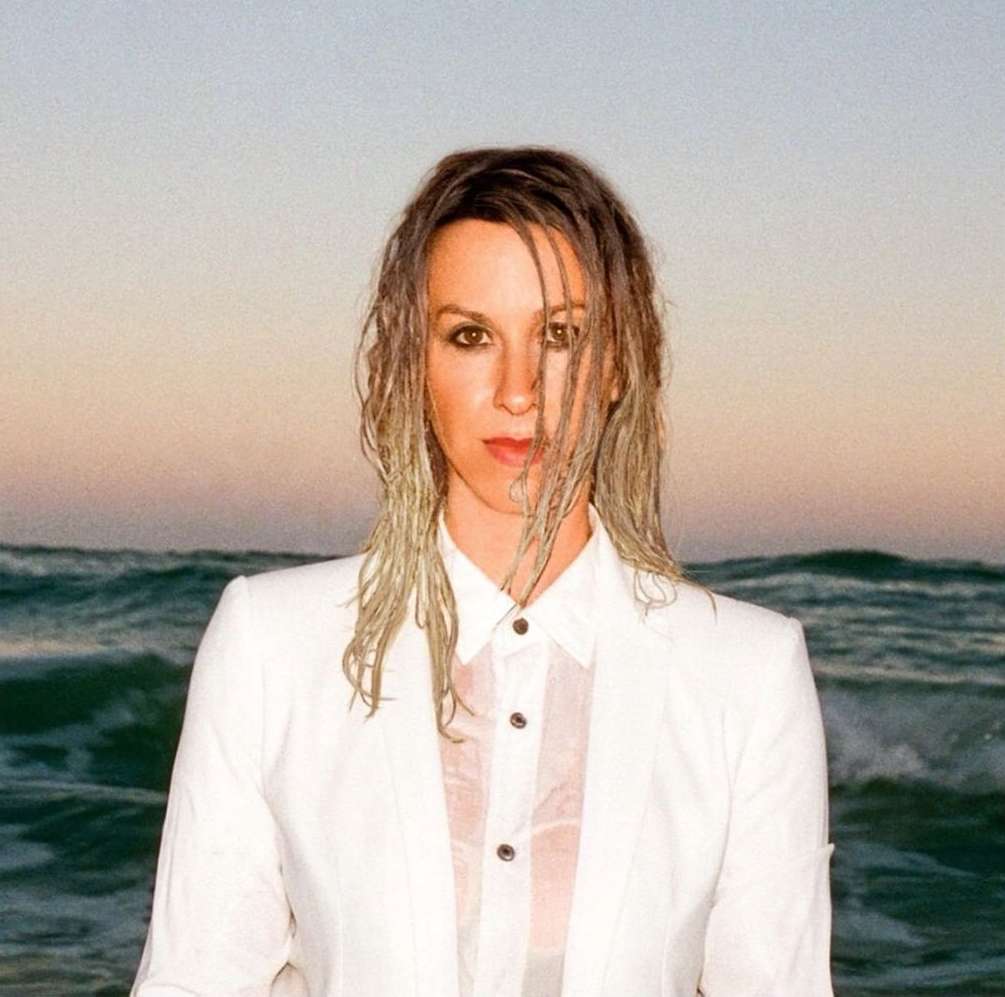 Woman with wet hair and white suit poses in front of ocean waves at sunset for a portrait shot with an eye-level angle.