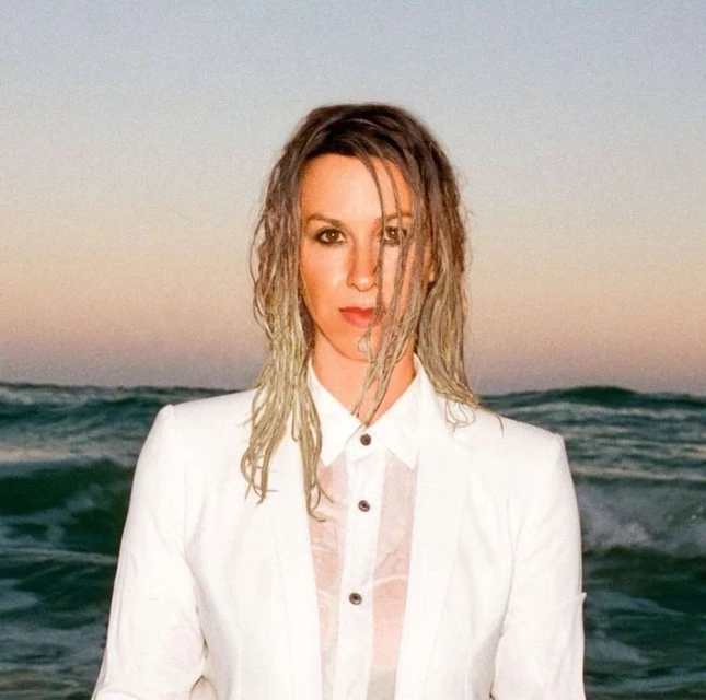 Woman with wet hair and white suit poses in front of ocean waves at sunset for a portrait shot with an eye-level angle.