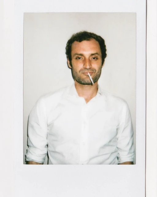 Indoor portrait of an adult male smoking a cigarette, wearing a white button-down shirt against a neutral white background.