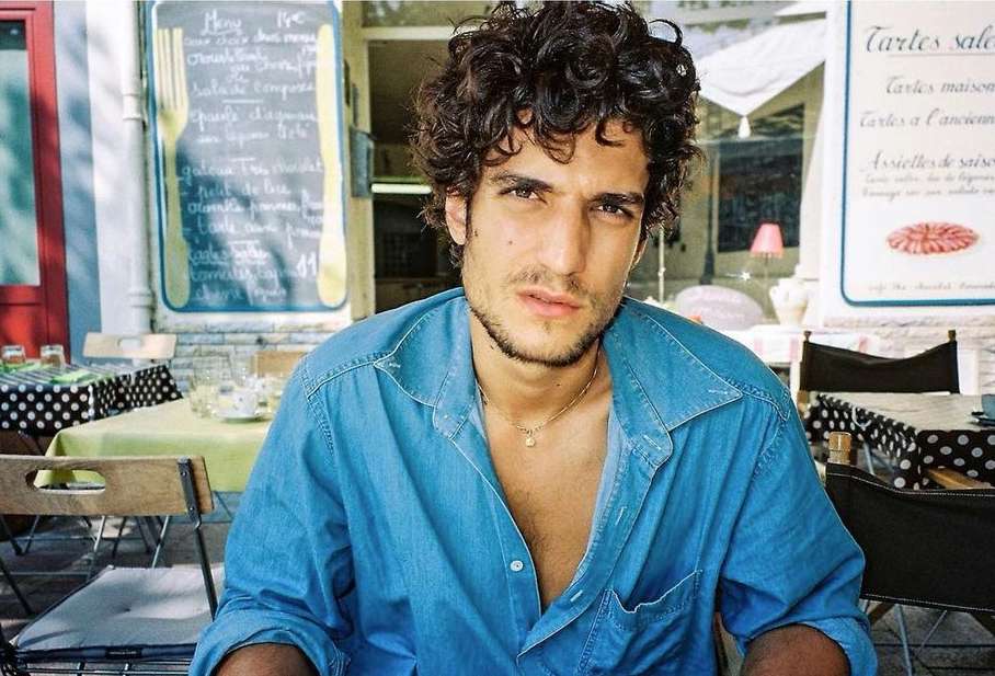 Close-up of a curly-haired man in a blue denim shirt sitting at an outdoor cafe with tables and chairs under a canopy.
