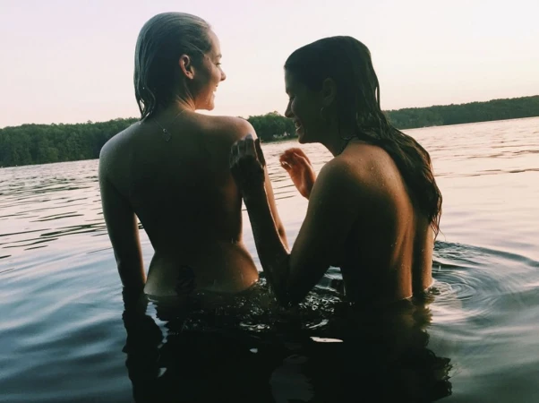 Two young women with wet hair sitting waist-deep in a lake at sunset facing each other and laughing.