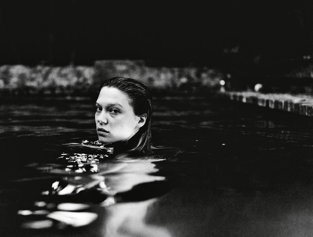 Black and white portrait of a woman in dark water, looking toward the camera with wet hair and neutral expression, with dark background.