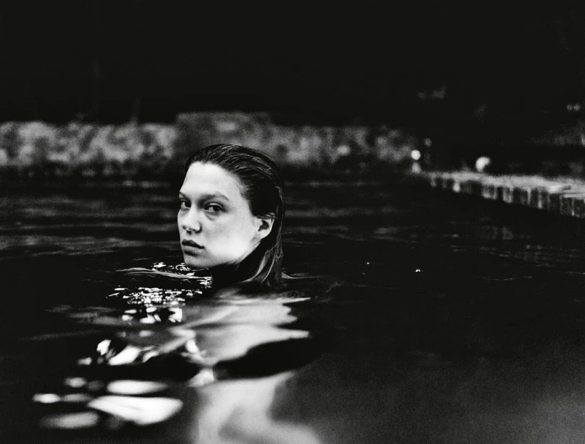 Black and white portrait of a woman in dark water, looking toward the camera with wet hair and neutral expression, with dark background.