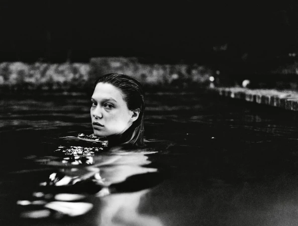 Black and white portrait of a woman in dark water, looking toward the camera with wet hair and neutral expression, with dark background.