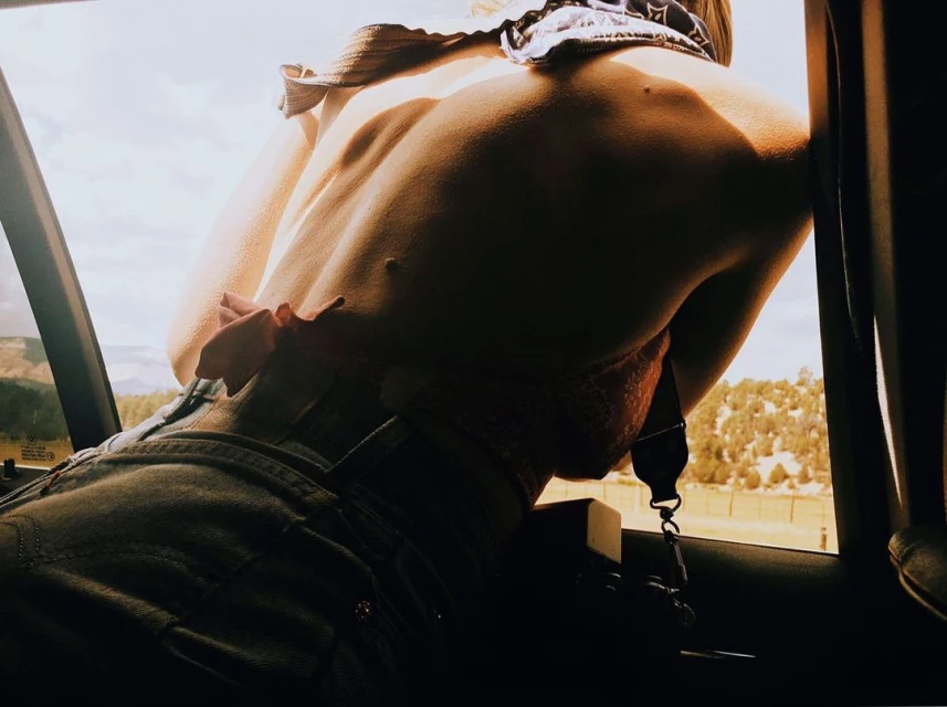 Back view of person looking out a car window at a sunny forest landscape, wearing blue jeans, a pink bra, and a bandana.